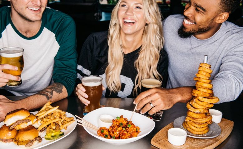 Three people sitting and laughing at a table full of food and beer